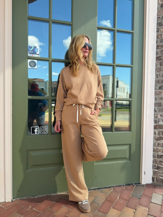 A woman wearing a matching beige sweatshirt and wide-leg sweatpants lounge set stands in front of an olive green glass door, with brick flooring and a brick wall visible.