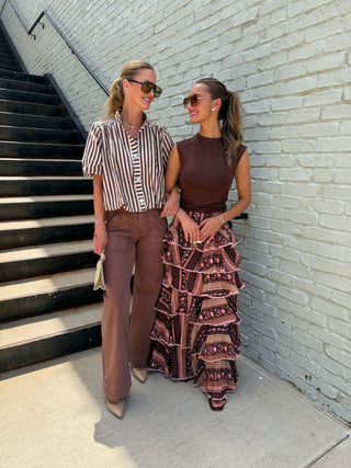 Two women standing outdoors next to a white brick wall and black stairs, wearing coordinated outfits. One wears a brown and white striped short-sleeve button-up shirt with brown pants, and the other wears a dark brown sleeveless top with a long tiered skirt in a pink, brown, and black pattern.