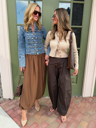 Two women standing outdoors in front of a green door, wearing coordinated outfits. The woman on the left wears a light blue denim jacket with silver buttons, a brown maxi skirt, beige heels, and a leopard-print handbag.