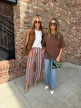 Two women posing outdoors in front of a brick wall. The woman on the left wears a brown leather jacket, white top, and red-and-white striped wide-leg pants.